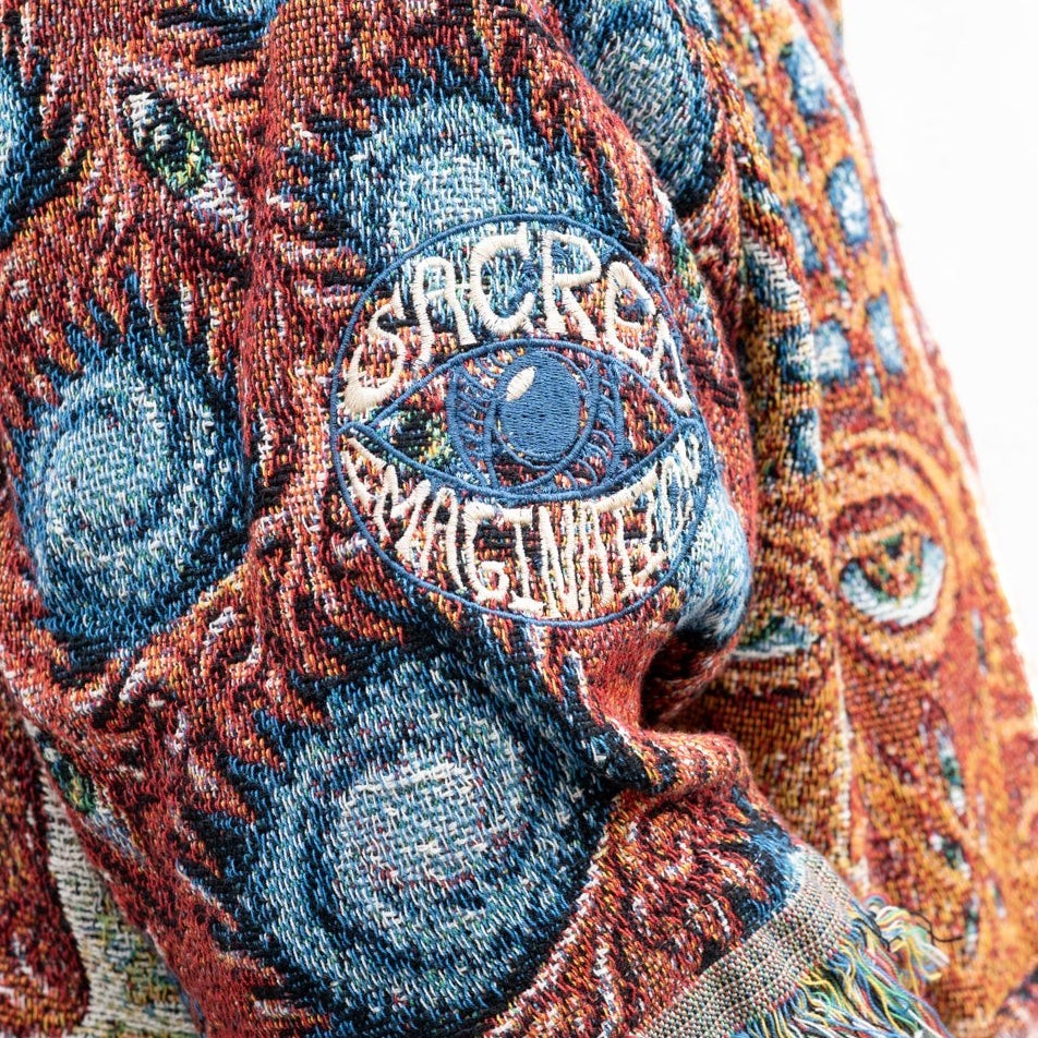 CoSM Shop Godself woven coat with intricate blue, orange, and red tapestry pattern and embroidered Sacred Imagination eye logo on sleeve.