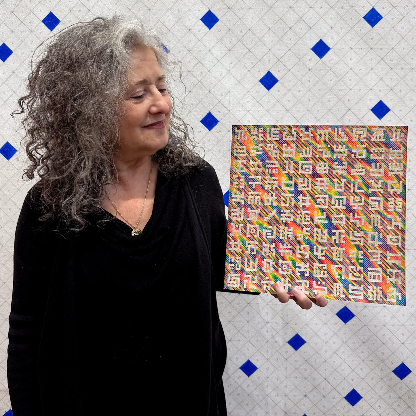 Allyson Grey holds a 12x12 inch pearlescent paper print of her painting "Pearlescent Secret Writing in Spectral Rain" featuring rainbow colors and unique symbols.