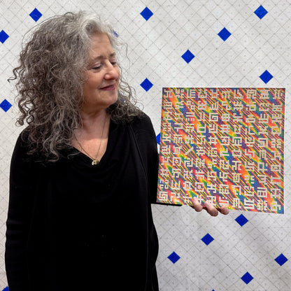 Allyson Grey holds a 12x12 inch pearlescent paper print of her painting "Pearlescent Secret Writing in Spectral Rain" featuring rainbow colors and unique symbols.