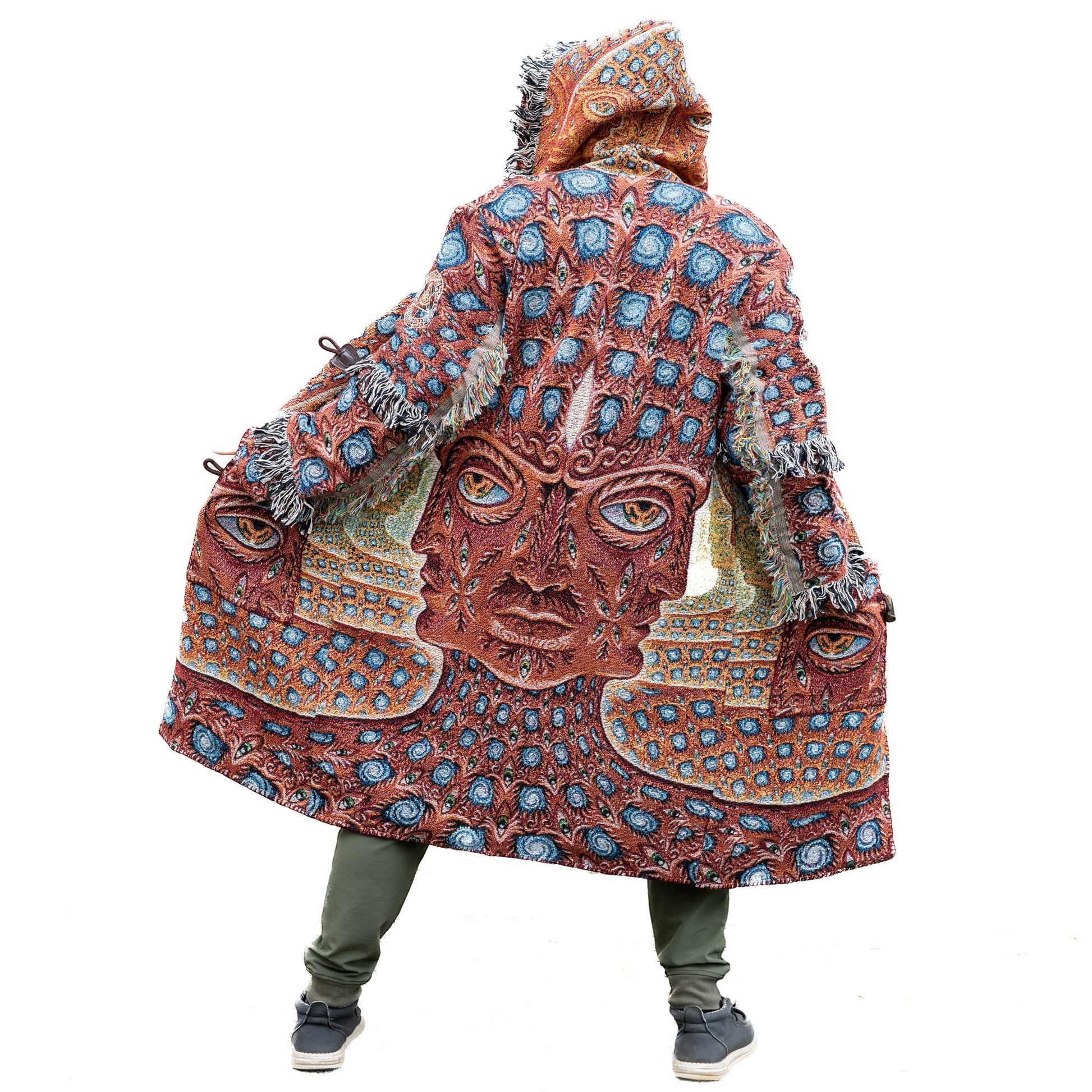 CoSM Shop unisex one-size woven coat with a hooded design and fringe trim, featuring a vibrant, intricate pattern of faces and eyes in earthy tones and blue.