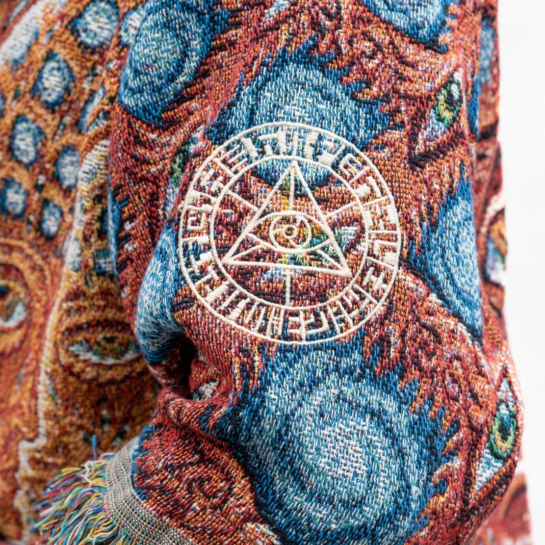 CoSM Shop unisex one-size woven coat with intricate eye and geometric patterns in blue, orange, and tan. Features embroidered emblem on sleeve.